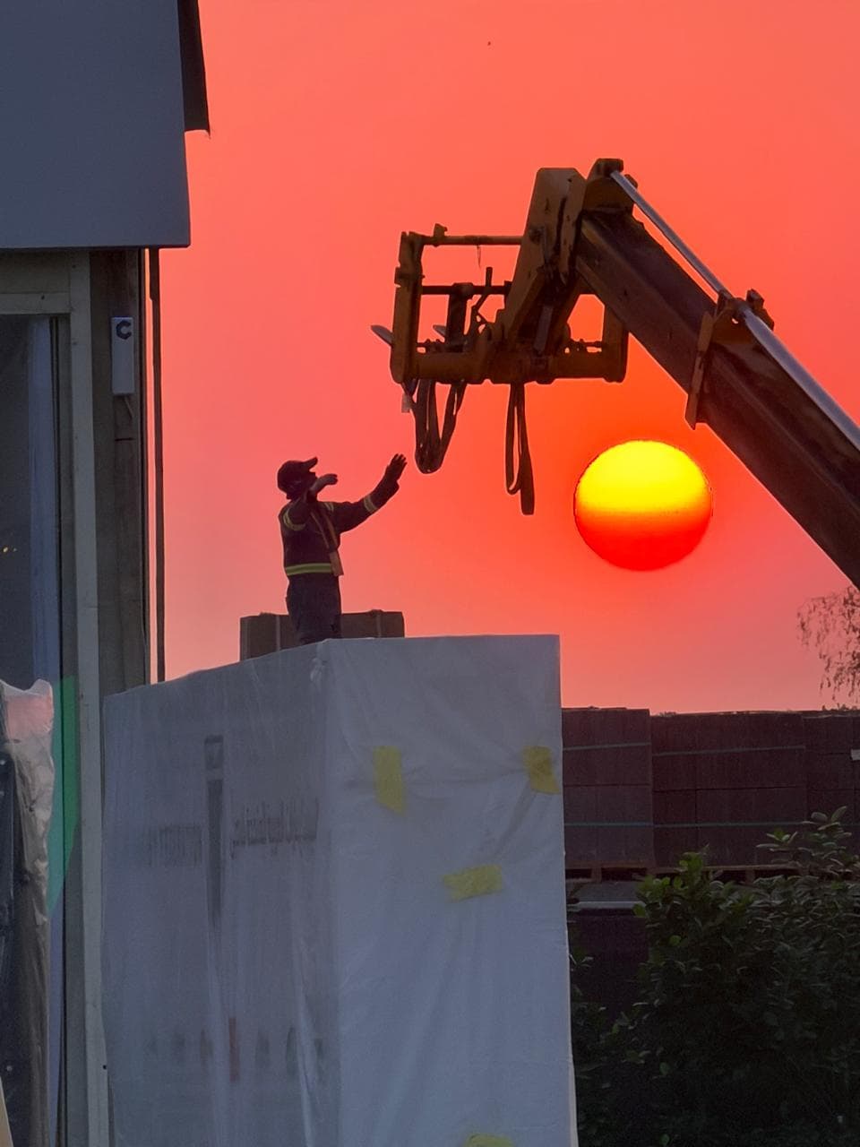 TotalEnergies build at sunset — a telehandler lifts a wrapped load as a worker signals from the structure