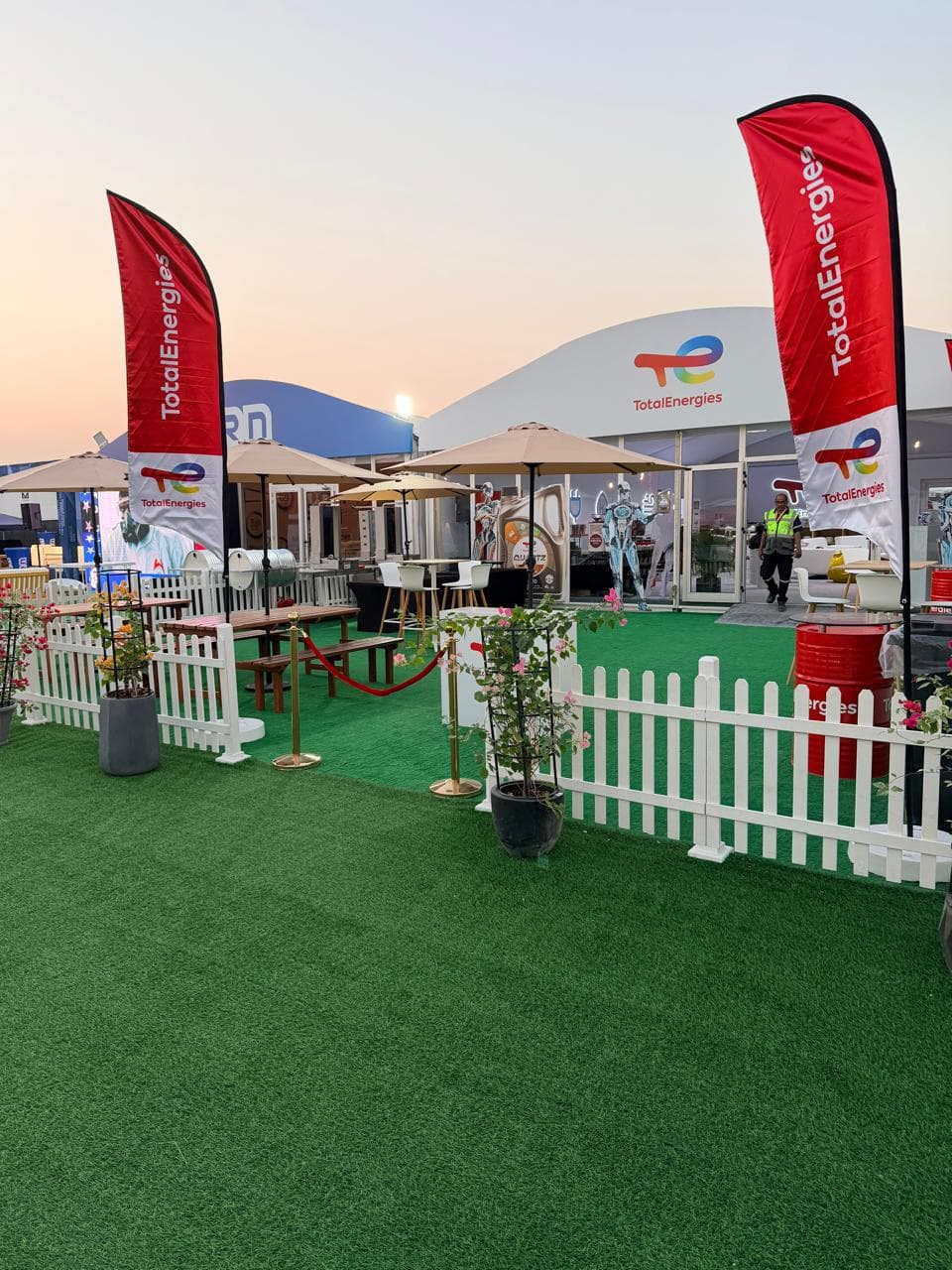 TotalEnergies hospitality tent at dusk with branded red feather flags, picket-fence perimeter and umbrella seating