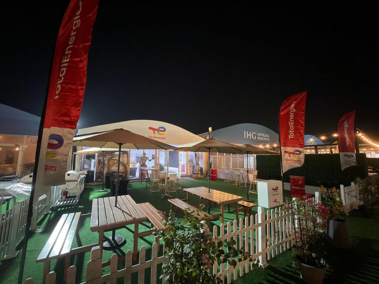 TotalEnergies activation at night — feather flags, string lighting and seating beside the IHG pavilion